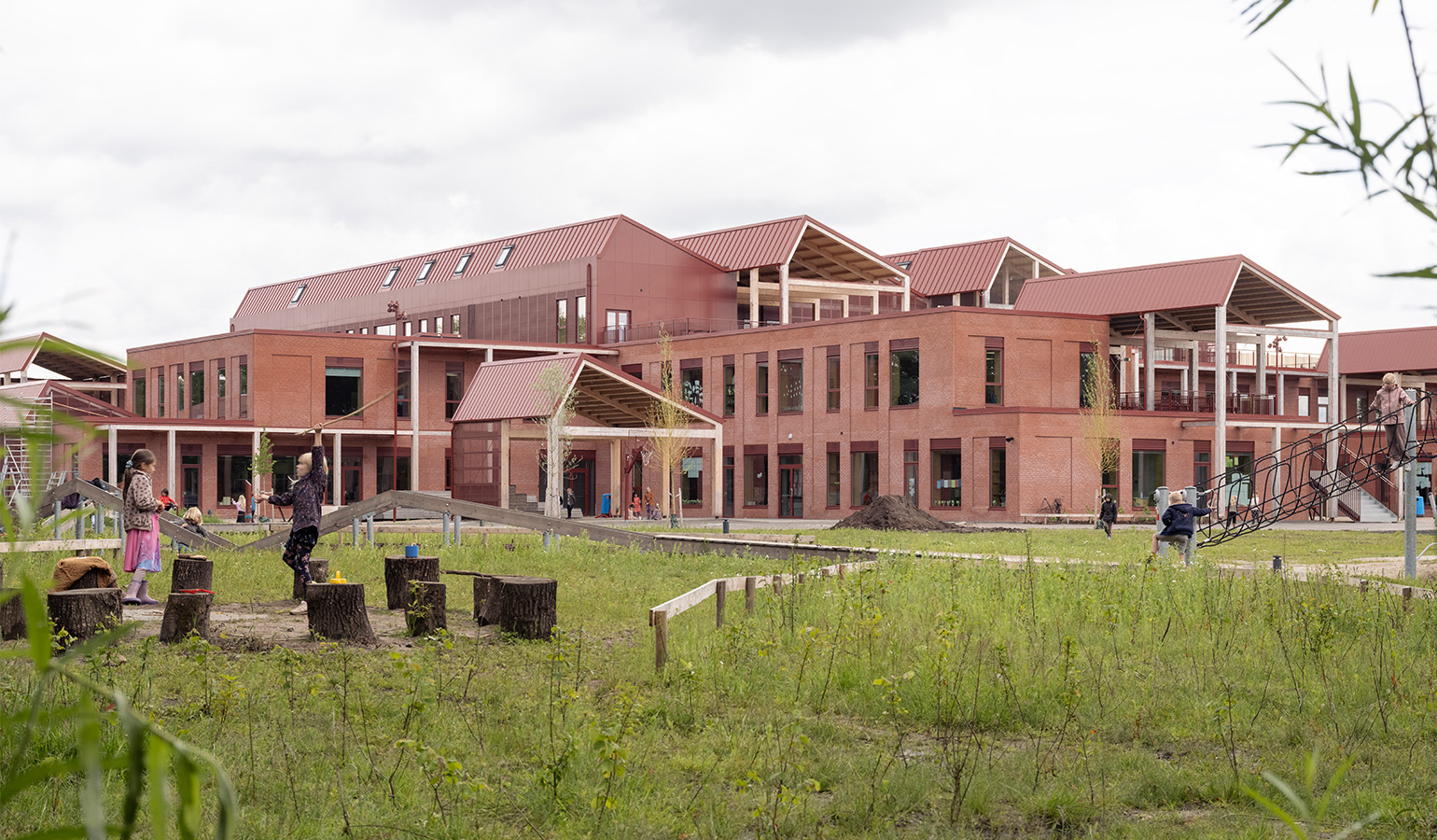 Side view of a red brick building in different sections with sloping roofs. In the foreground, children are playing in a green area.
