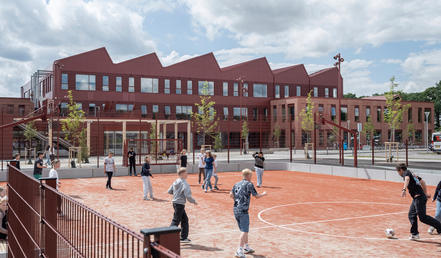 In the background, a school building in red brick with a roof made of sloping sections creating a zigzag pattern. In the foreground, a sports field.