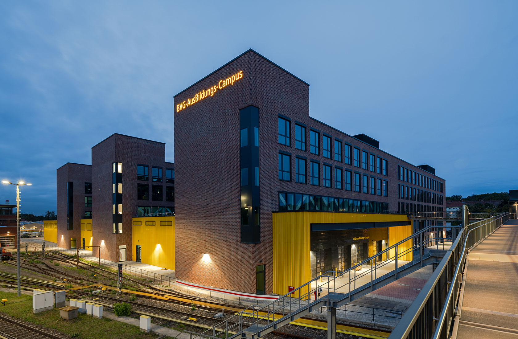 Three red brick buildings are placed parallel to each other, with lower yellow buildings between them that connect the buildings to one another. A railway yard to the left of the building.