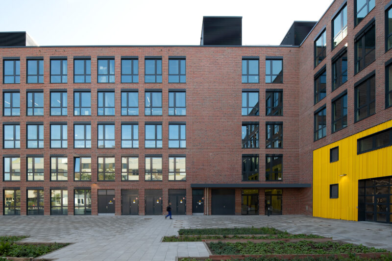 Building with five floors made of red brick with large window areas. Gray paving in front of the building.
