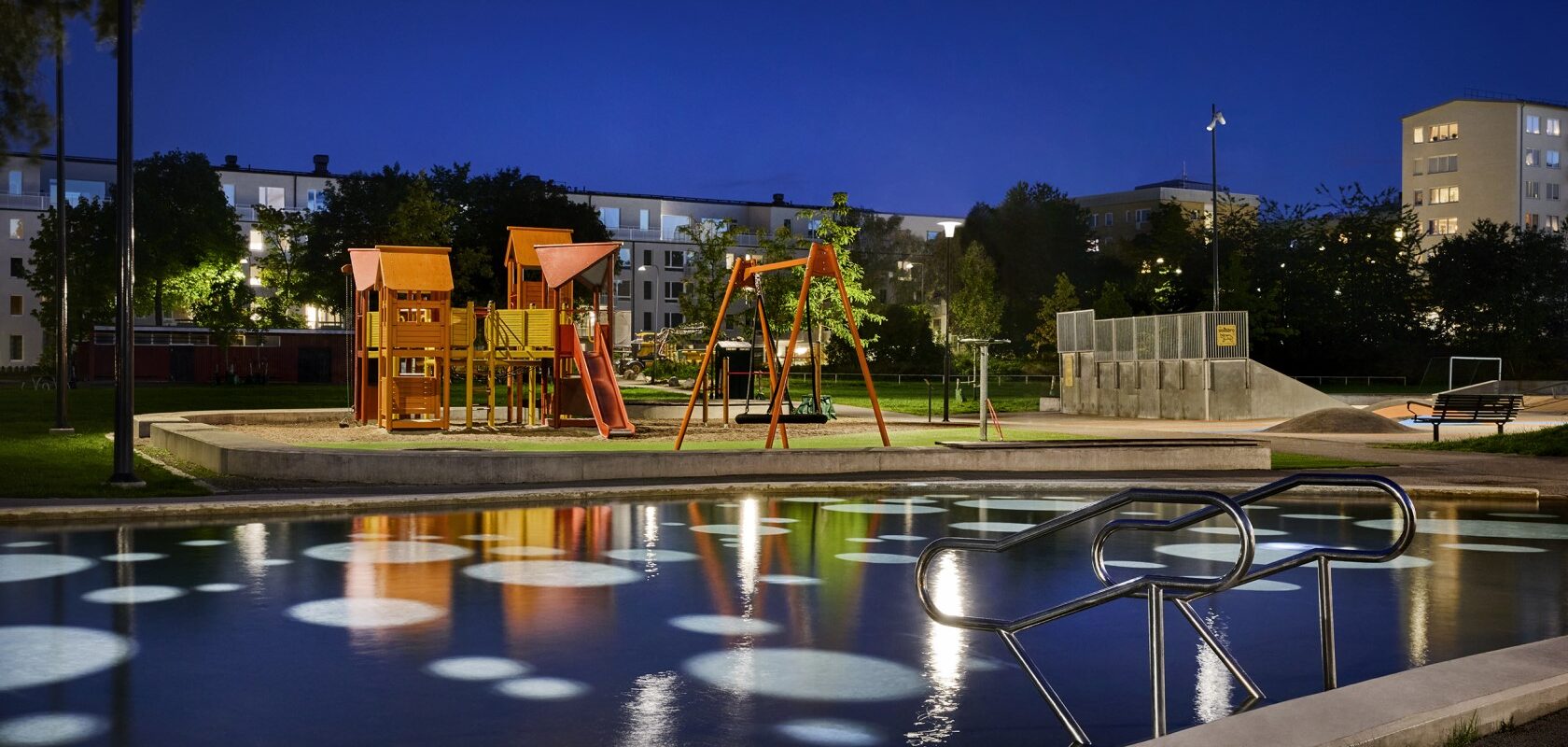 Evening image of a larger illuminated playground with a pond in front. In the background, there are trees and residential buildings.
