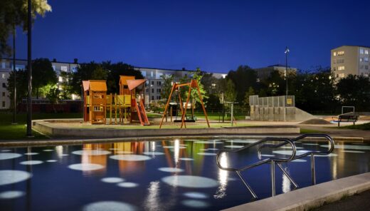 Evening image of a larger illuminated playground with a pond in front. In the background, there are trees and residential buildings.