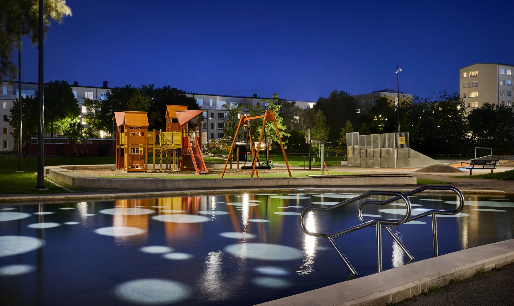 Evening image of a larger illuminated playground with a pond in front. In the background, there are trees and residential buildings.