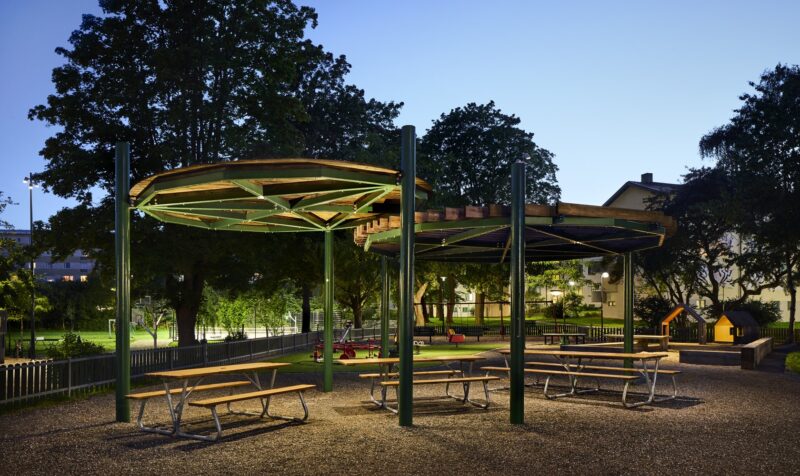 Park with three tables with benches under a roof, nicely lit in the evening.