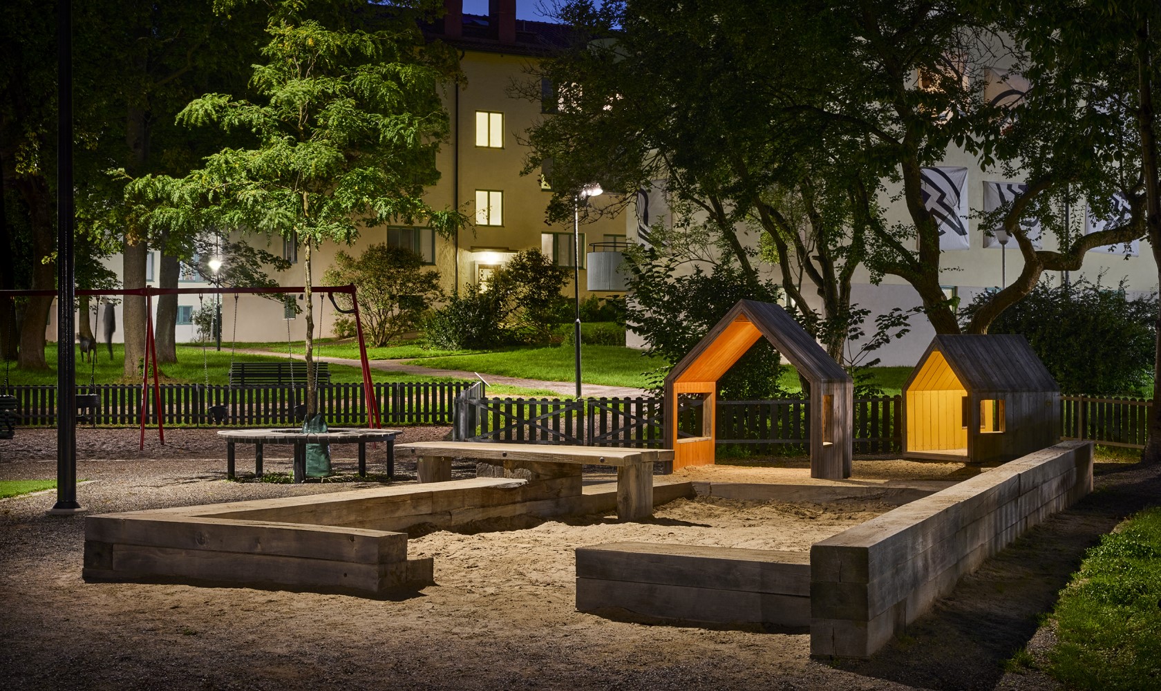 Playground by a green area with a sandpit, small illuminated houses, and residential buildings in the background.