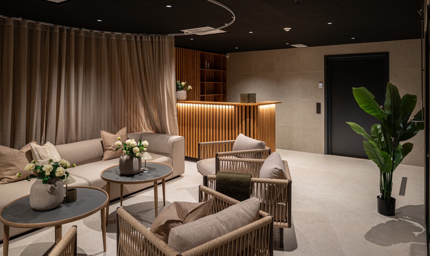 Part of a relaxation area with a seating group and armchairs partially made of rattan, next to the wall on the right a smaller wooden reception desk.