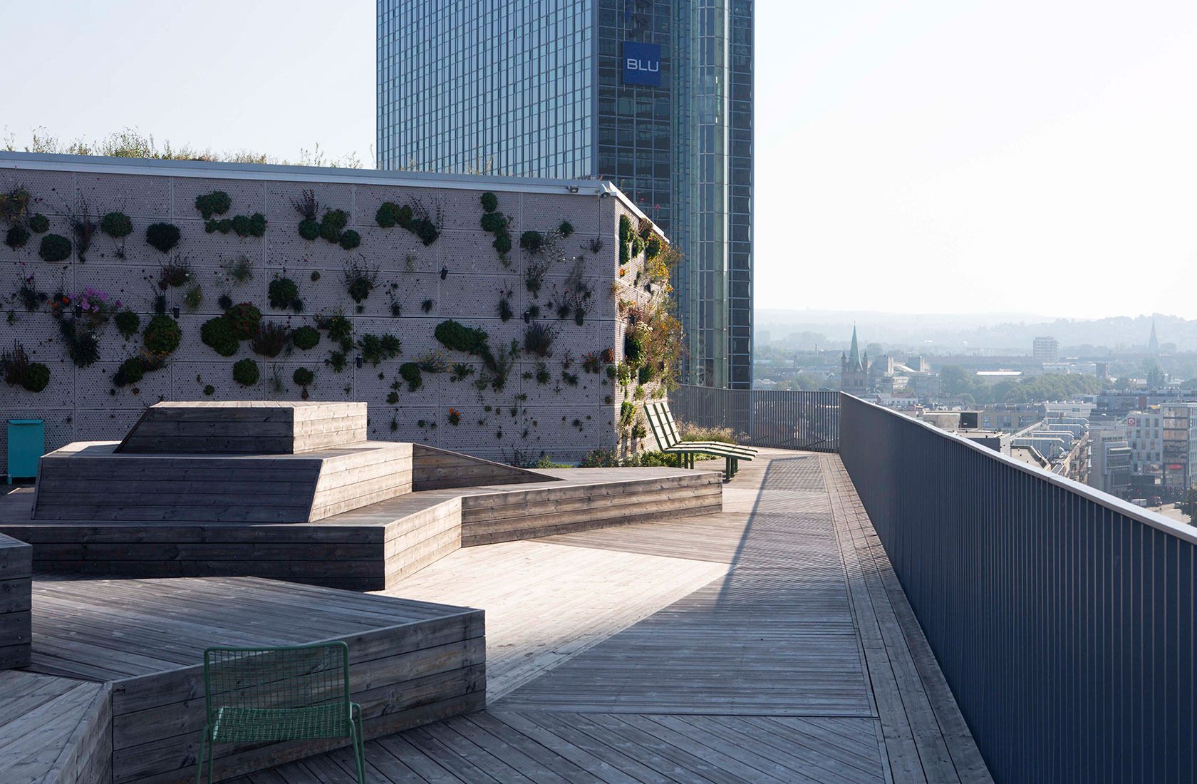 Wooden rooftop terrace with a white facade that is partially covered with plants, various types of seating areas with a view over the rooftops in central Oslo.