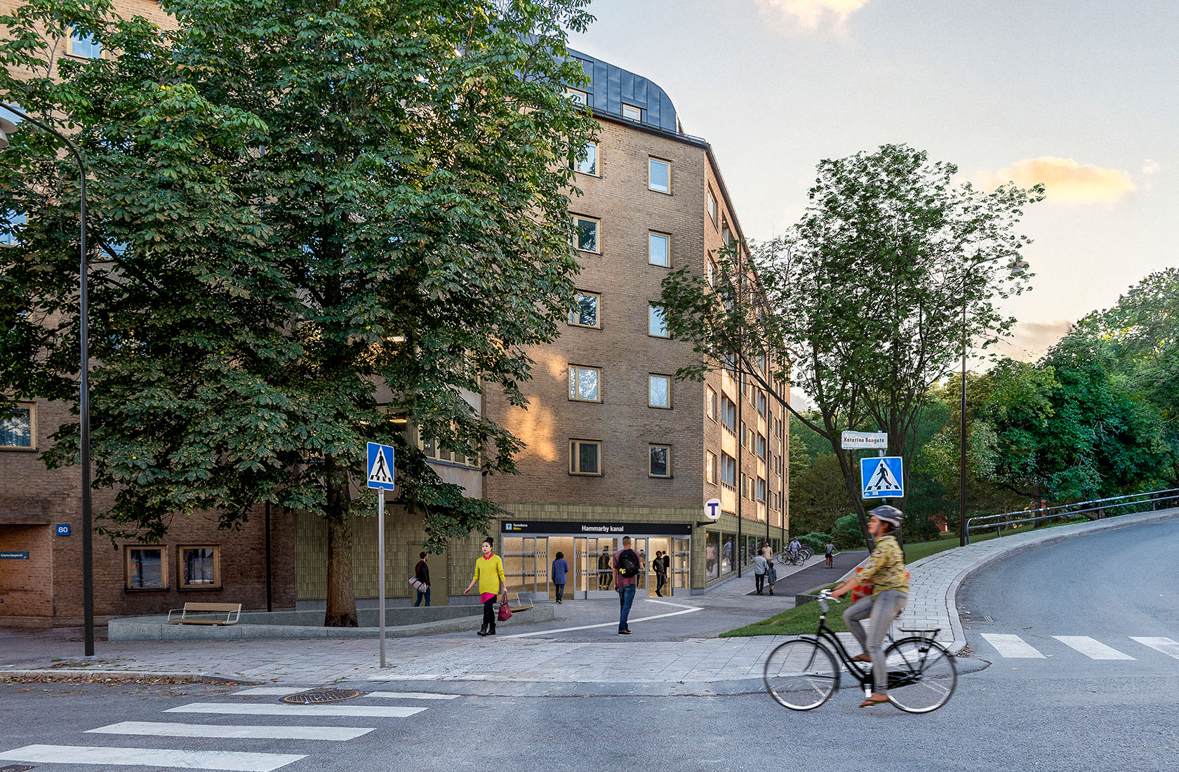 Visualization of the entrance to the subway station connected to a residential building, with a road and a cyclist in front, and a green area to the right.