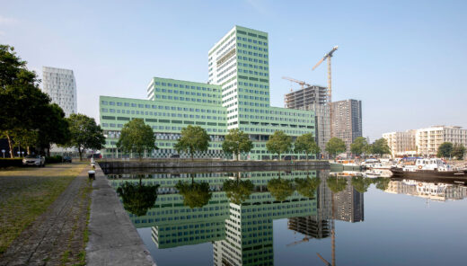 Large hospital building in green, where the building consists of blocks with varying heights by a harbor area with smaller boats in the foreground.