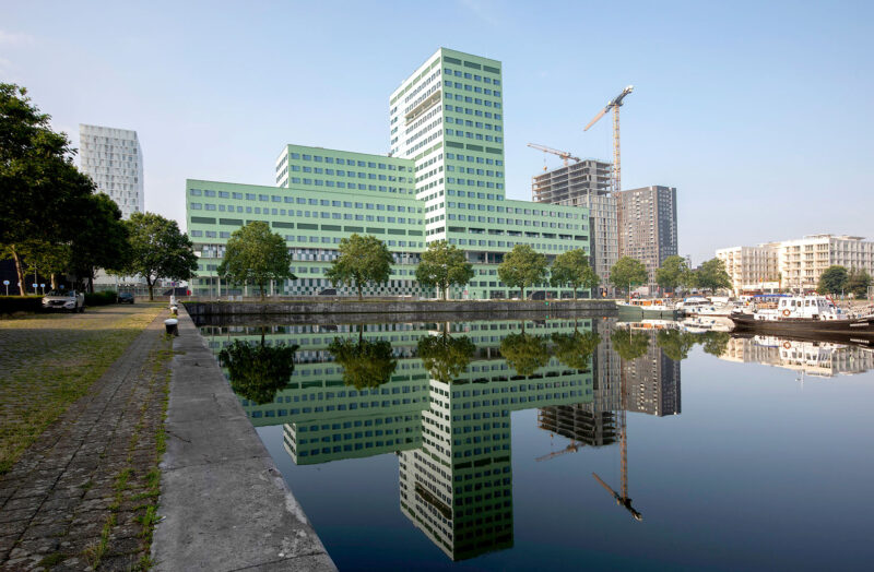 Large hospital building in green, where the building consists of blocks with varying heights by a harbor area with smaller boats in the foreground.