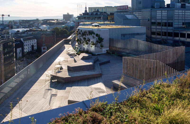 Image from above of a rooftop terrace with wooden flooring and elevated seating areas in wood, green roof surfaces, and some walls with vegetation.