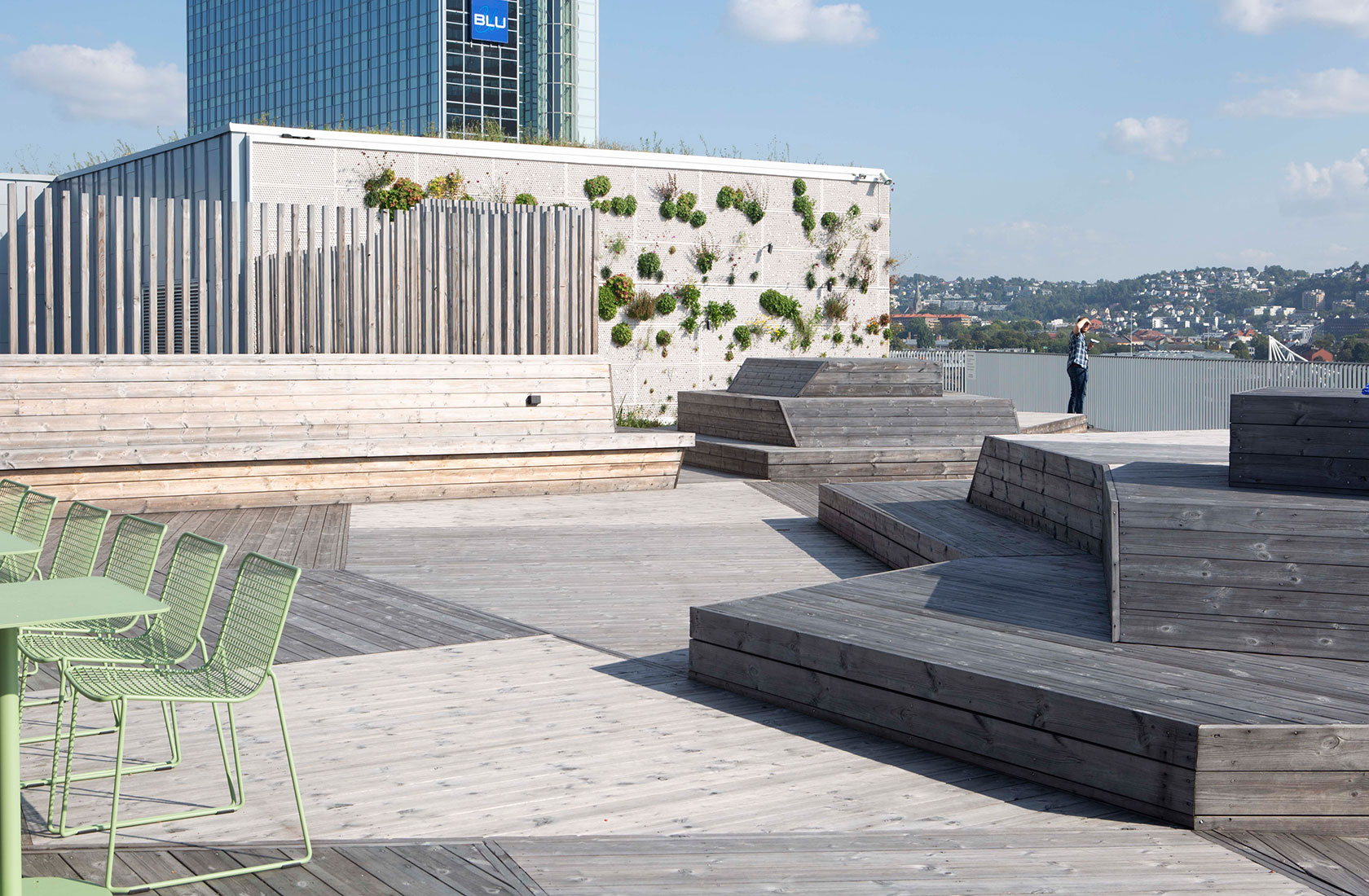 Wooden rooftop terrace with many uniquely shaped wooden seating areas, in the background a white wall that is partially covered with plants. Green chairs and tables to the left.