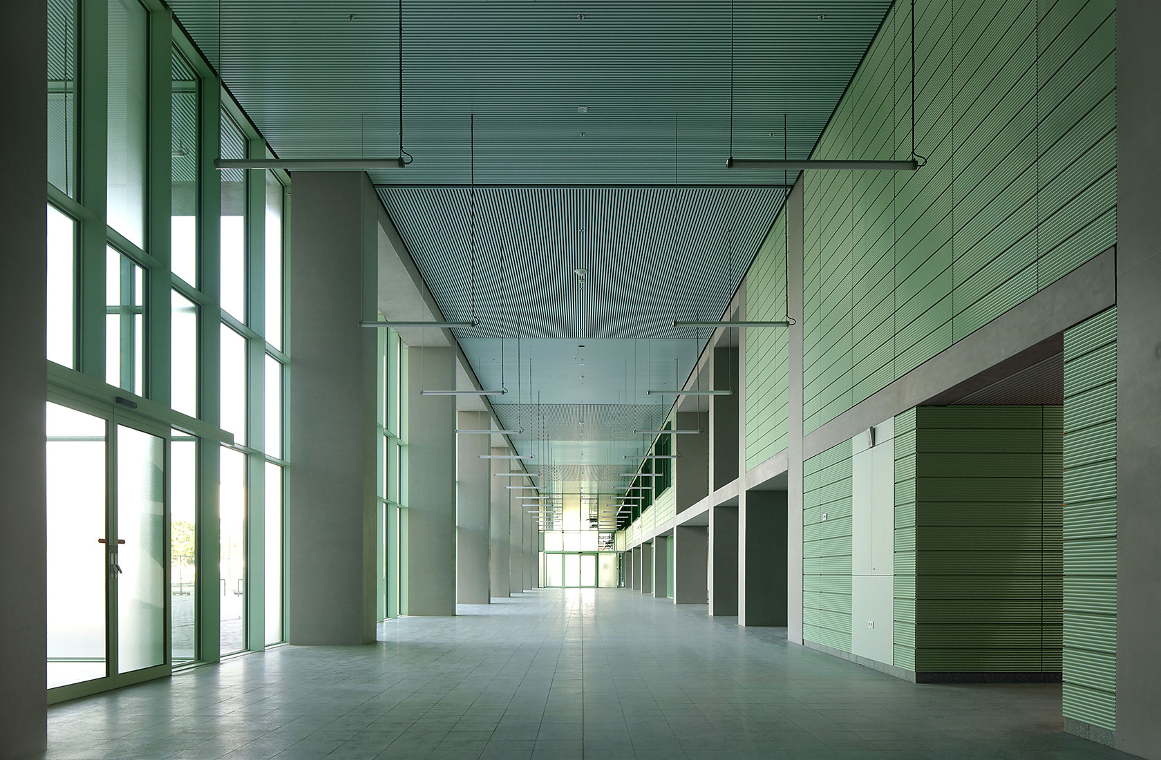 Empty corridor in the hospital building, wide and with a high ceiling, green wall on the right and glass section on the left.