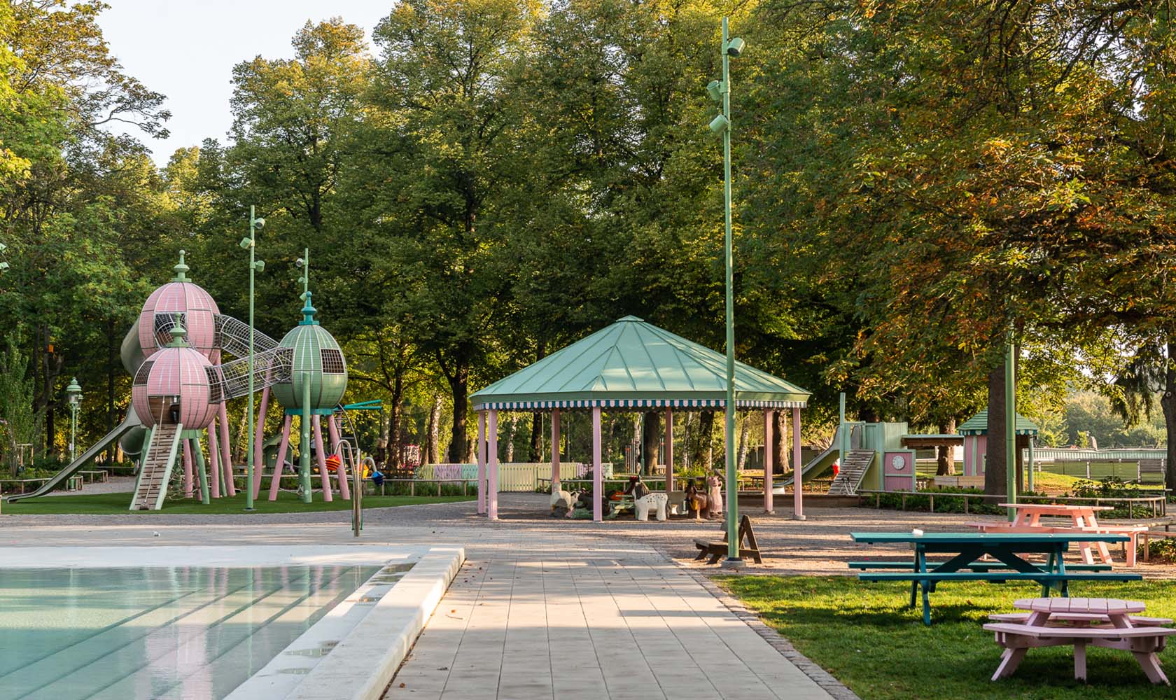 Tivoli-inspired playground area in a green park with a pool to the left and seating areas to the right.