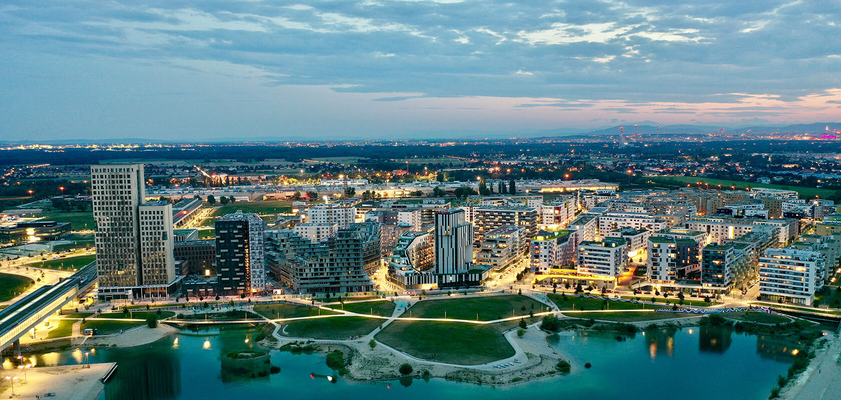 Aerial view of a modern cityscape with illuminated buildings, park, and water feature at dusk.