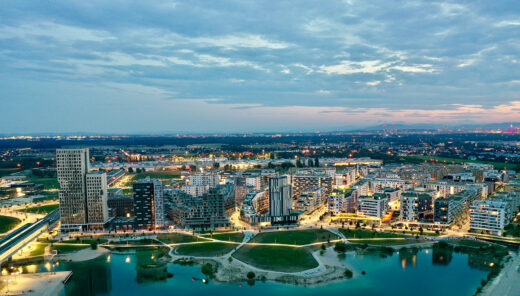 Aerial view of a modern cityscape with illuminated buildings, park, and water feature at dusk.