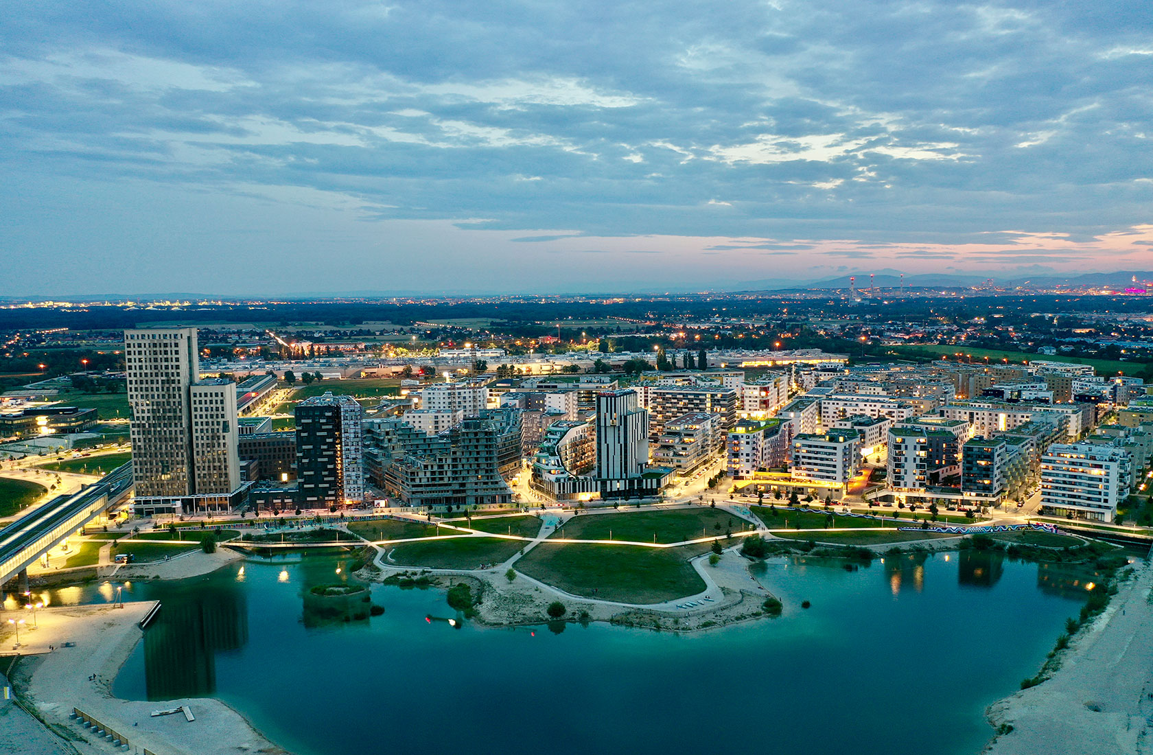 Aerial view of a modern cityscape with illuminated buildings, park, and water feature at dusk.