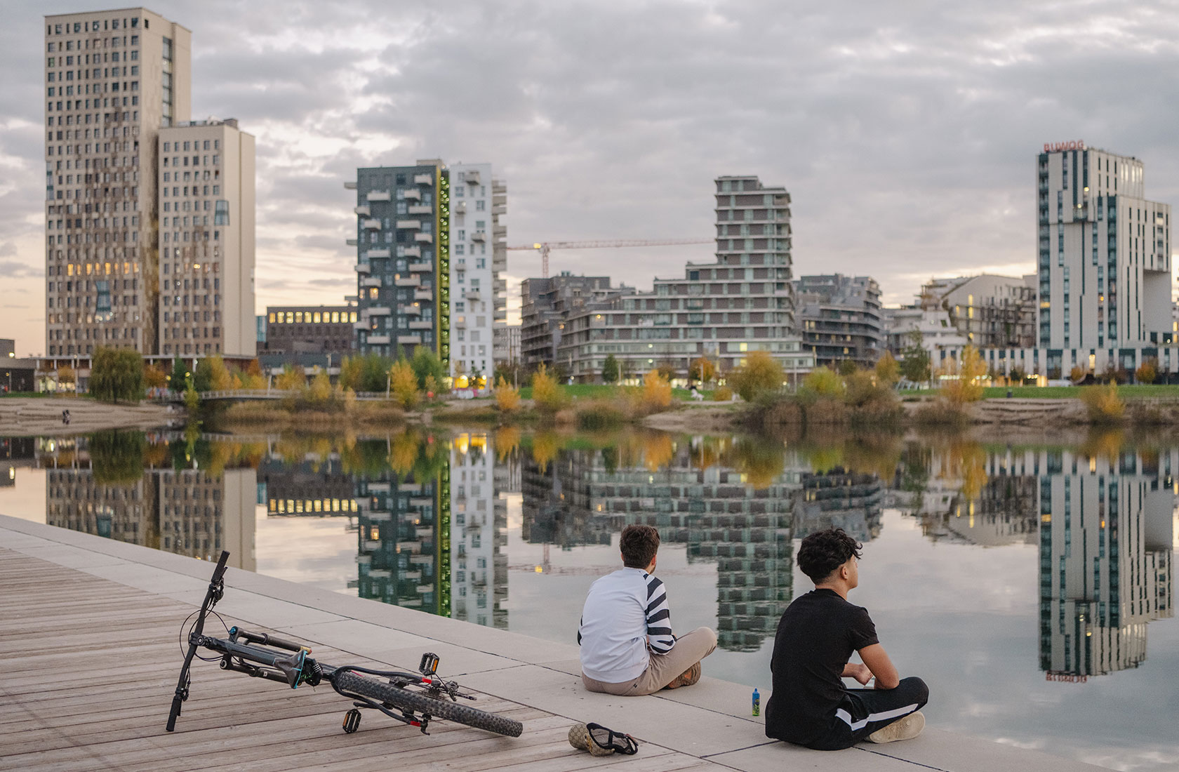 wo people sitting lakeside with bicycles, city skyline reflecting in the water at sunset.