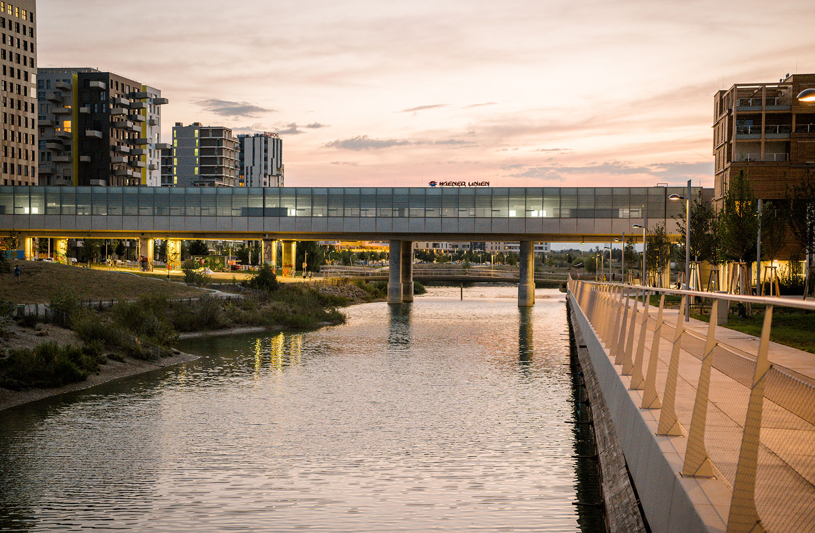 Modern pedestrian bridge over a canal at sunset, surrounded by contemporary buildings and greenery.