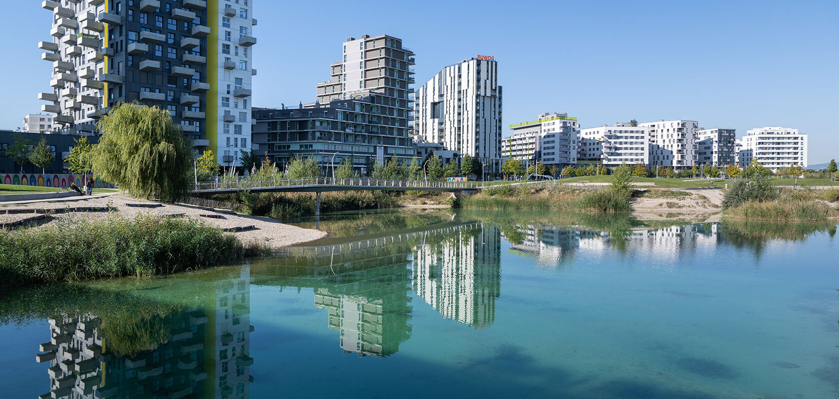 Modern residential buildings reflected in a clear lake under a bright blue sky.