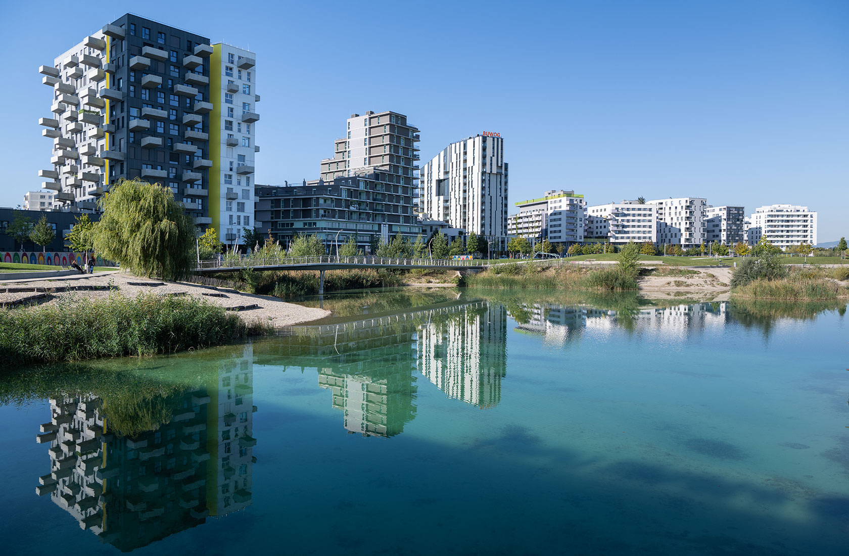 Modern residential buildings reflected in a clear lake under a bright blue sky.