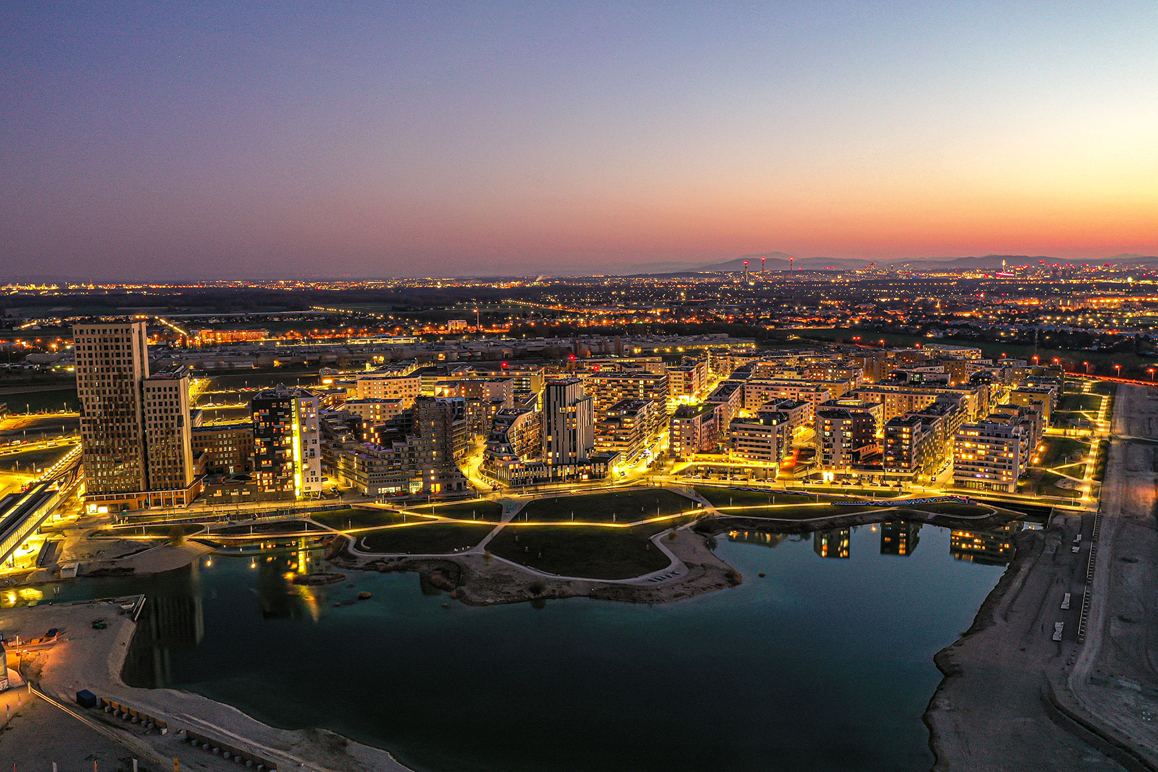 Aerial view of illuminated city buildings and lake at sunset with distant horizon.