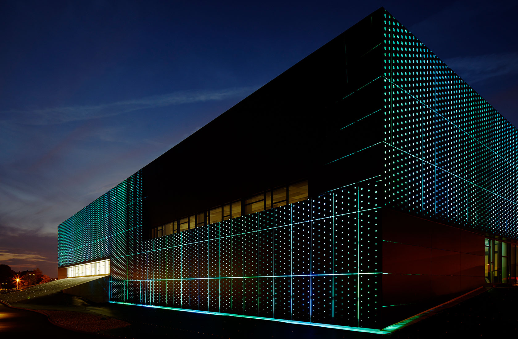 Evening view of the swimming hall with a facade in black metal with a perforated pattern and colored lighting behind it.