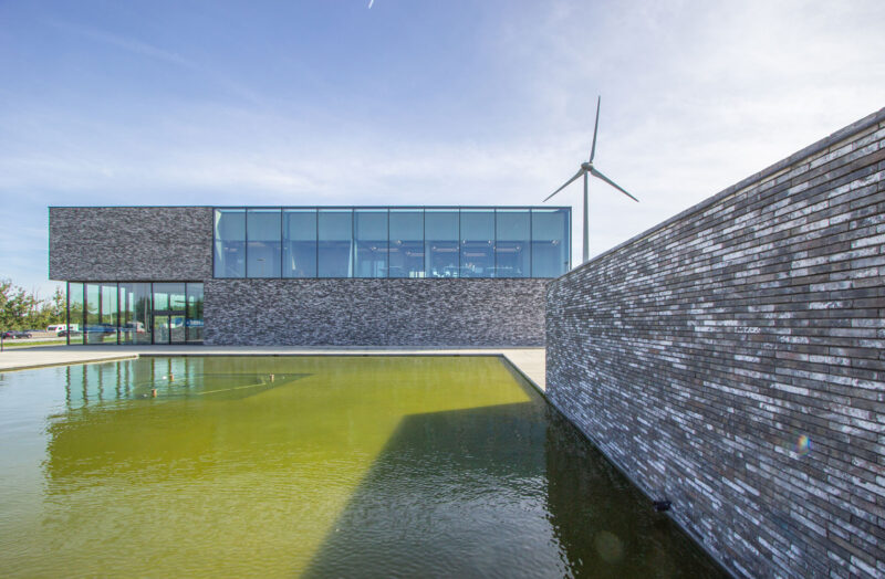 Modern building for robotic surgery with glass facade, wind turbine, and pond under clear sky.