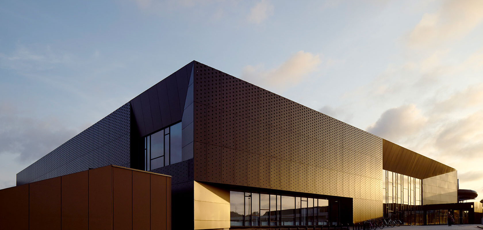 View from the side of a swimming hall made of black metal and large windows, where the sun gives it a golden shimmer in certain parts.