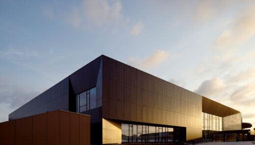 View from the side of a swimming hall made of black metal and large windows, where the sun gives it a golden shimmer in certain parts.