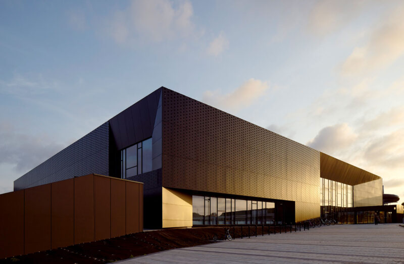 View from the side of a swimming hall made of black metal and large windows, where the sun gives it a golden shimmer in certain parts.