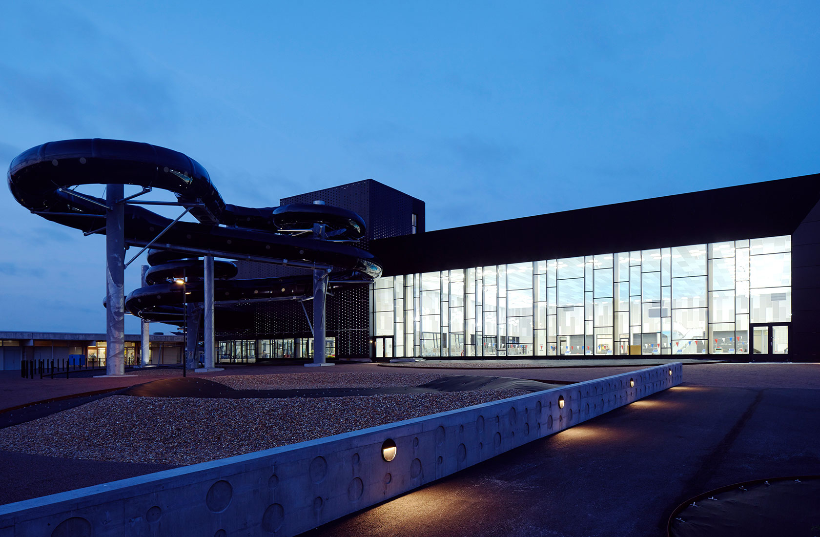 Evening view of the back of the swimming hall with a large window area at the pool section and an adventure area where a long water slide extends outside the building.