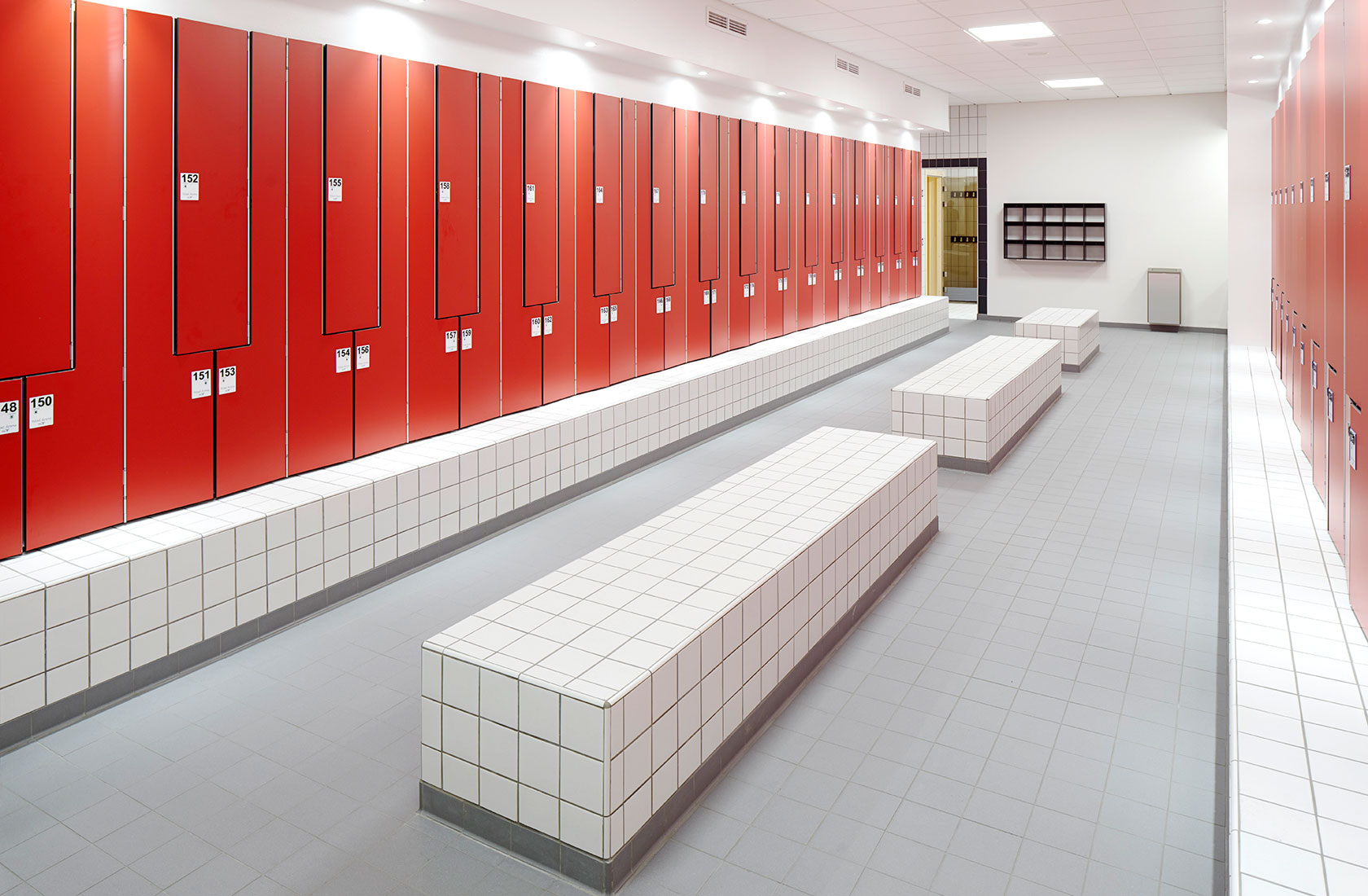 Changing room in the swimming hall with red locker doors and white tiles on the floor and benches.