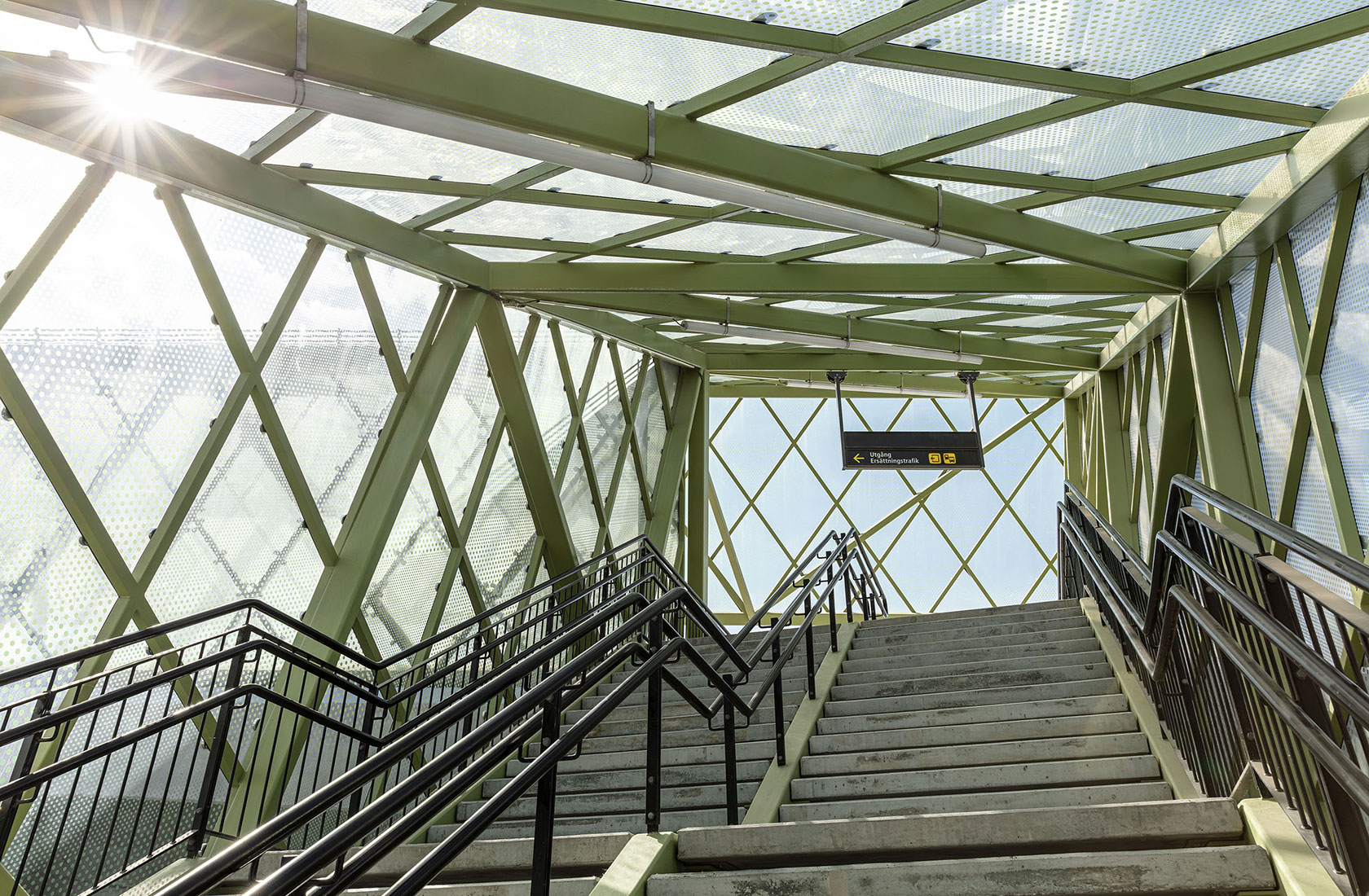 Modern glass and steel staircase from train platform with sunlight and sign for exit.