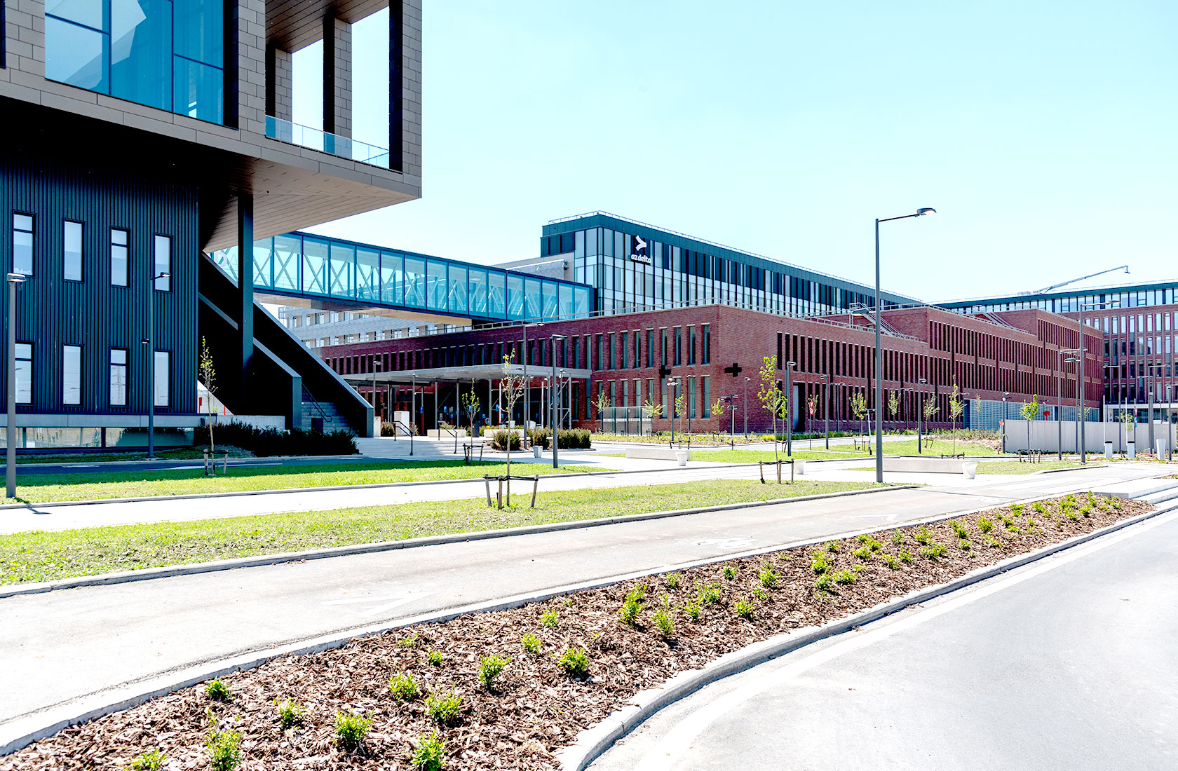 Modern hospital buildings with glass walkway, landscaped grounds, and clear blue sky.