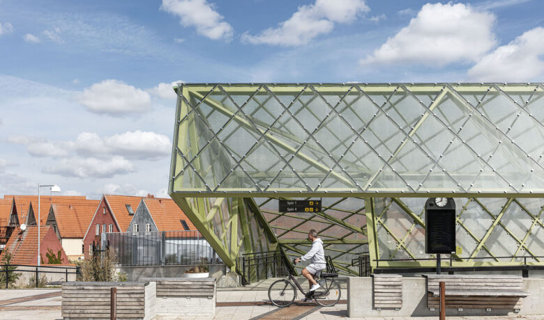 Man riding bike past modern train station with glass structure and houses under a blue sky with clouds.