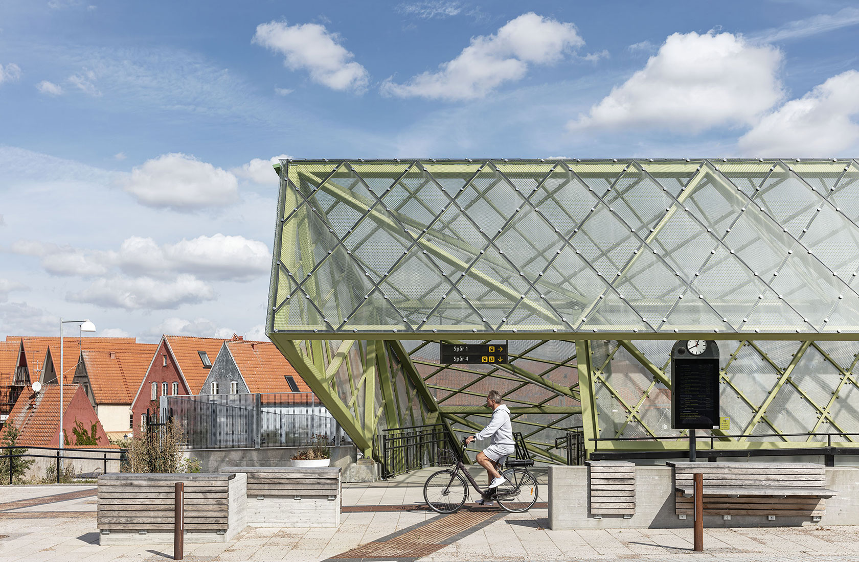Man riding bike past modern train station with glass structure and houses under a blue sky with clouds.