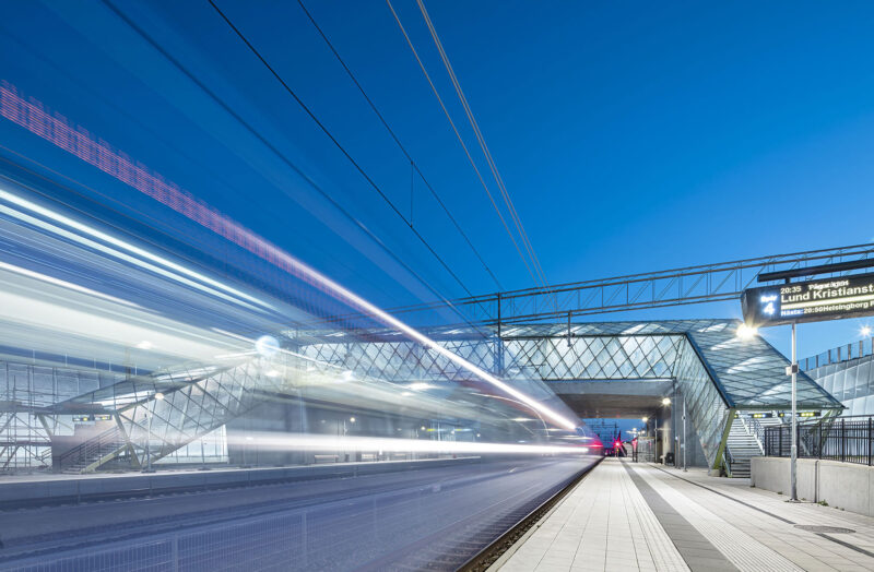 High-speed train passing through a modern station at night with bright lights and clear blue sky.