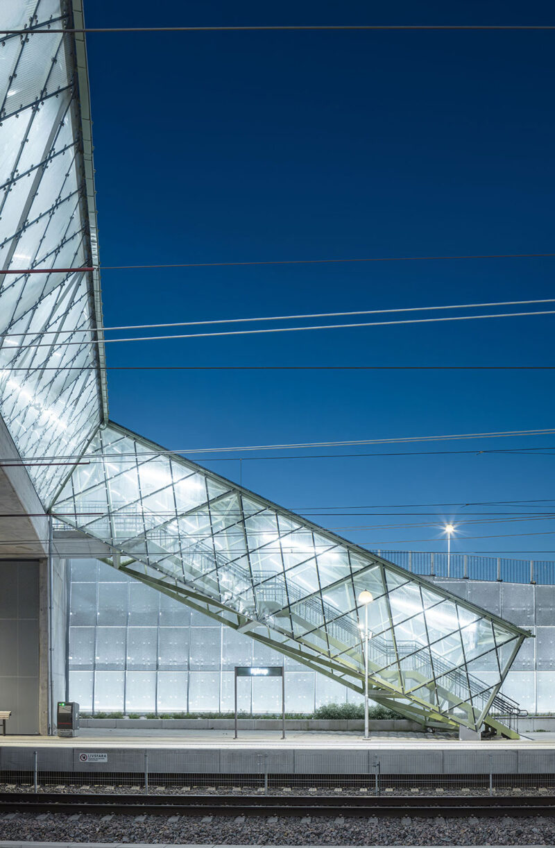 Modern glass staircase from train platform at night with illuminated interior and overhead power lines.