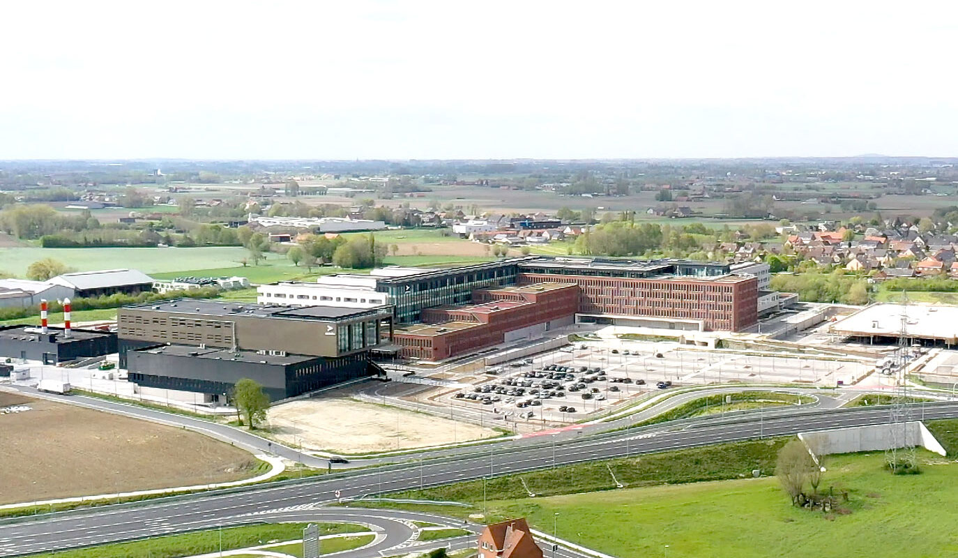 Aerial view of a modern hospital complex with parking lot, surrounded by fields and residential areas.