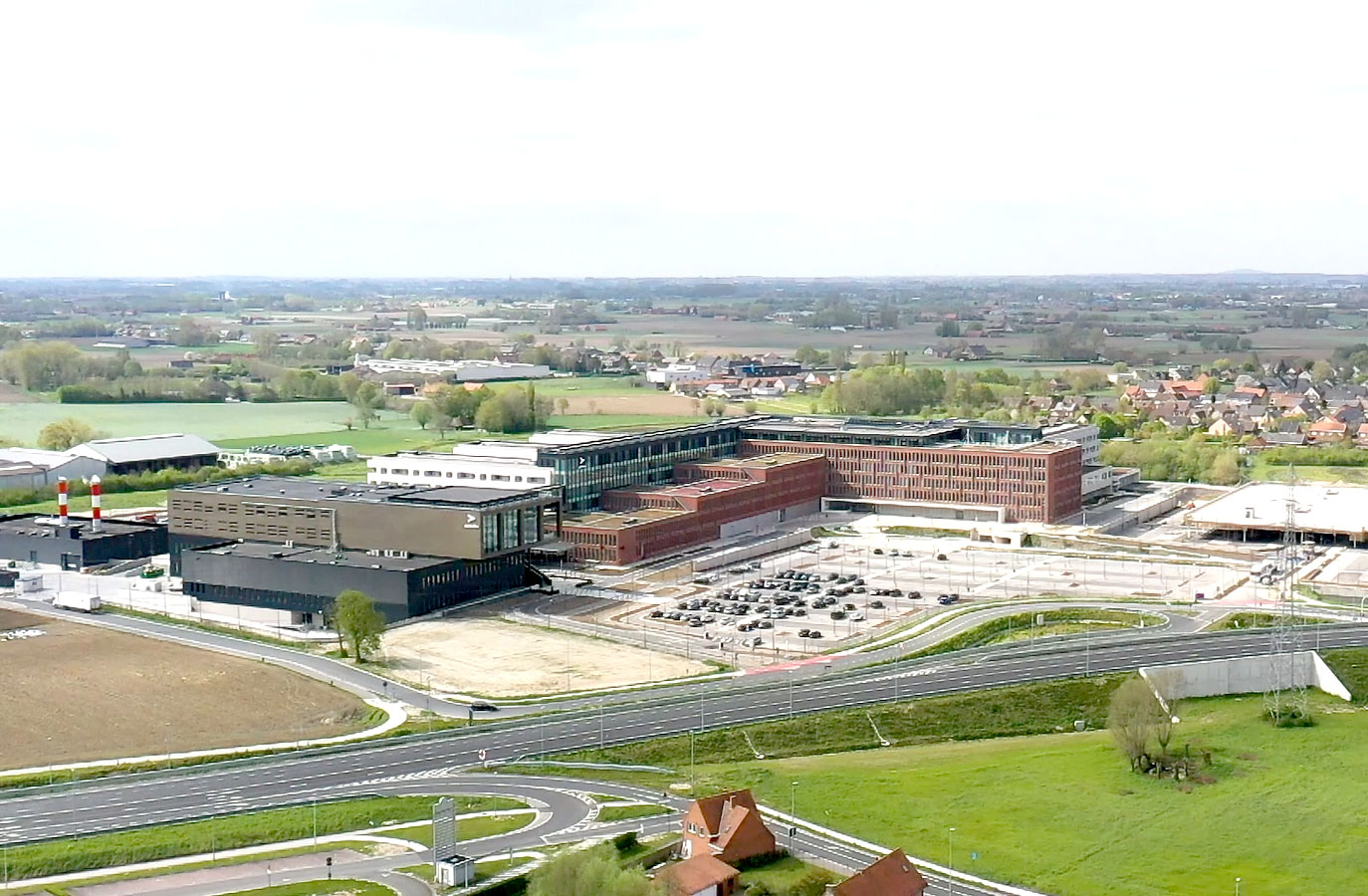Aerial view of a modern hospital complex with parking lot, surrounded by fields and residential areas.