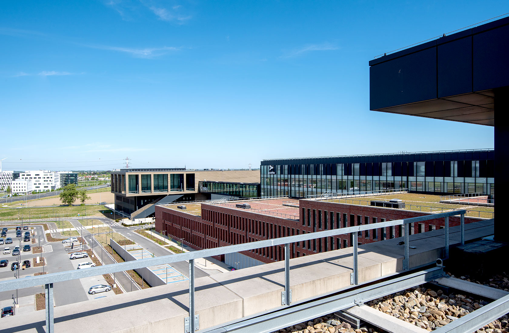 Modern hospital campus with brick and glass buildings under a clear blue sky, featuring parking area.