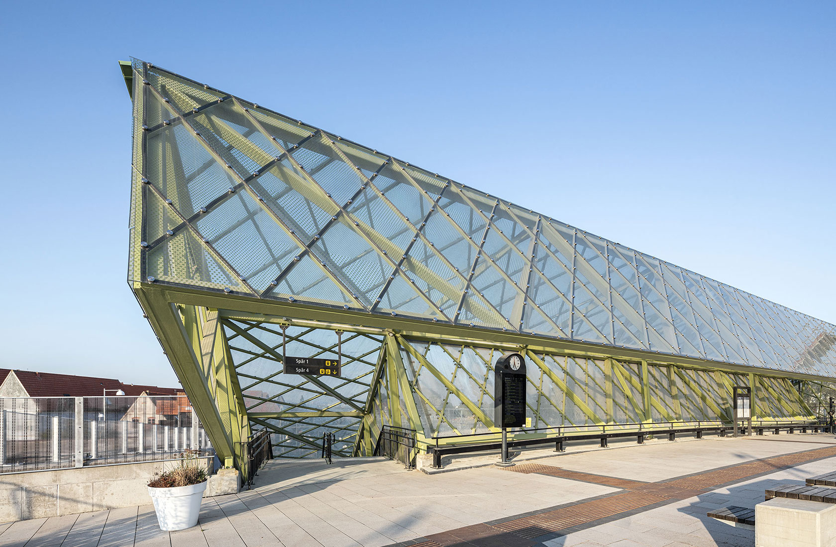 Modern glass and steel structure of train station with clear blue sky and building in background.