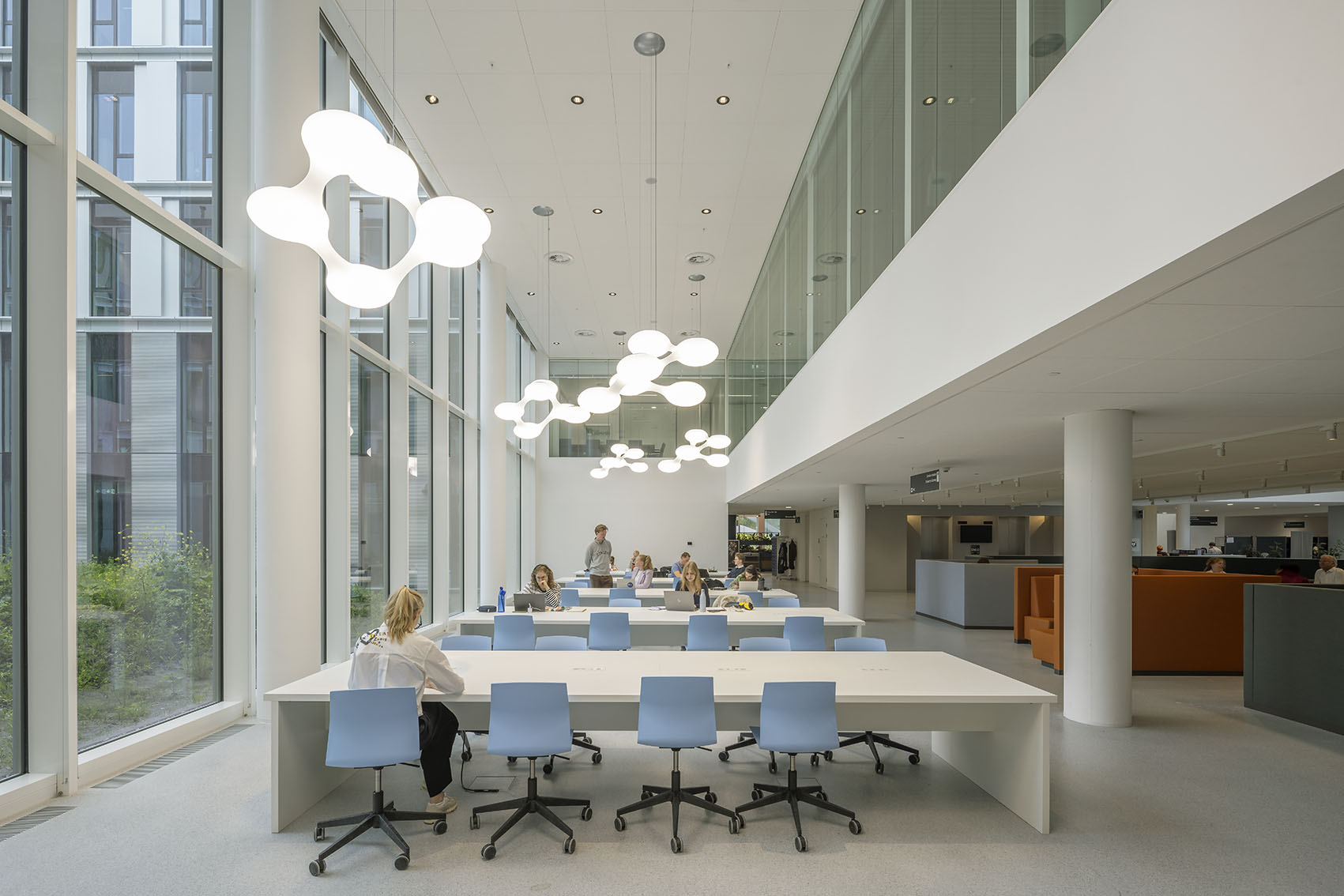 Modern office space with large windows, stylish lighting, and employees working at shared desks.