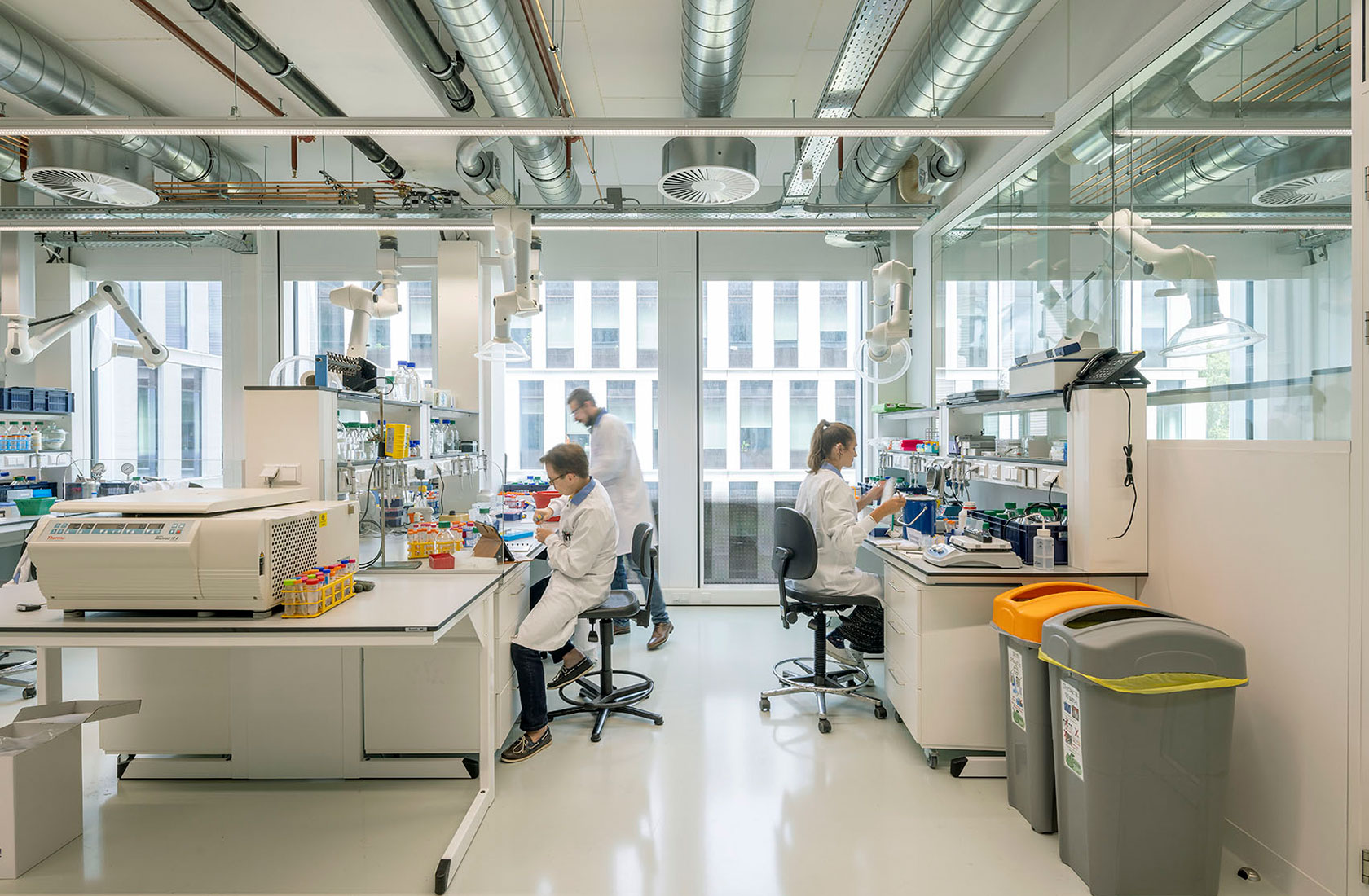 Scientists working in a modern laboratory with equipment and waste bins visible.
