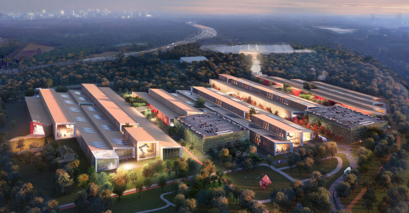 Aerial view of a modern industrial complex surrounded by trees with city skyline in the distance.