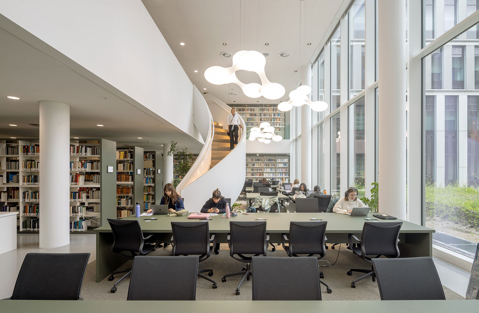 Modern library with large windows, students studying at tables, and a spiral staircase in the background.