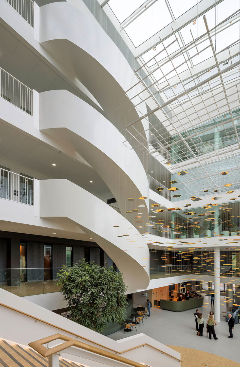 Modern office atrium with spiral stairs, hanging decor, large plant, and employees conversing.