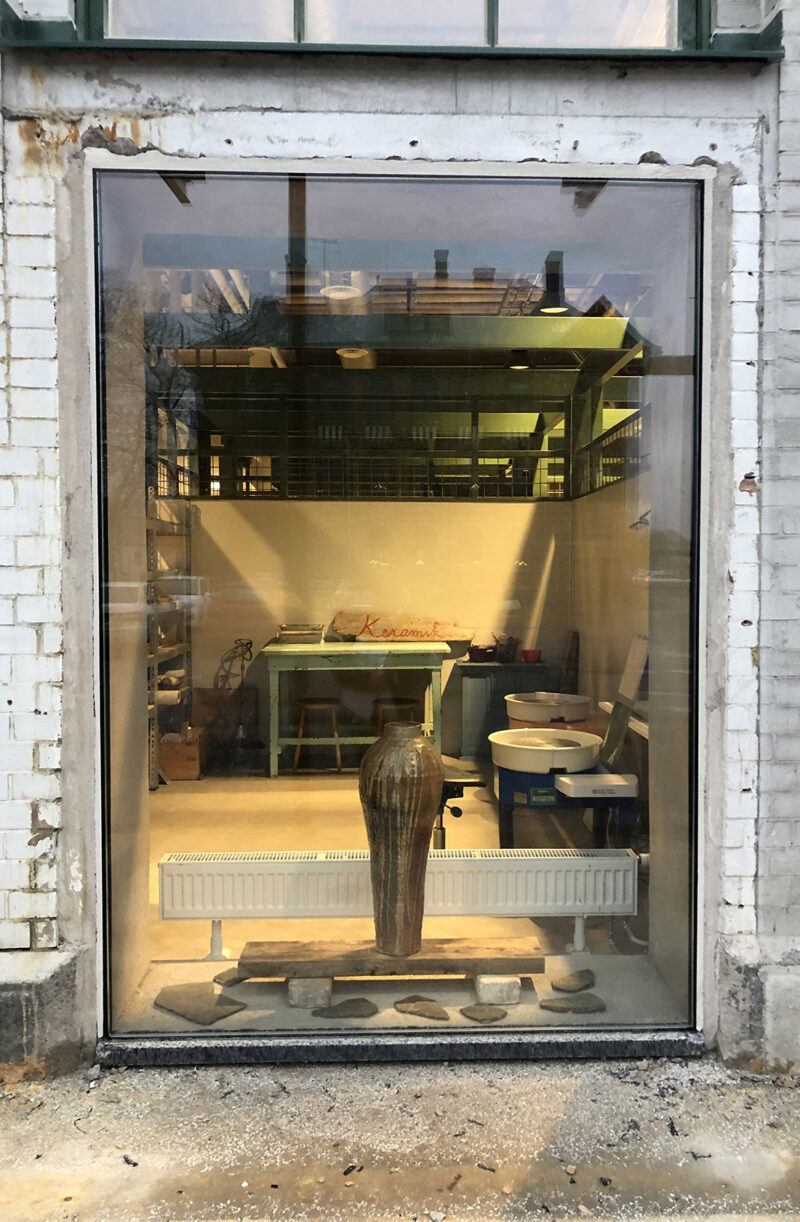 Rustic pottery studio with large window display featuring a ceramic vase and pottery equipment.