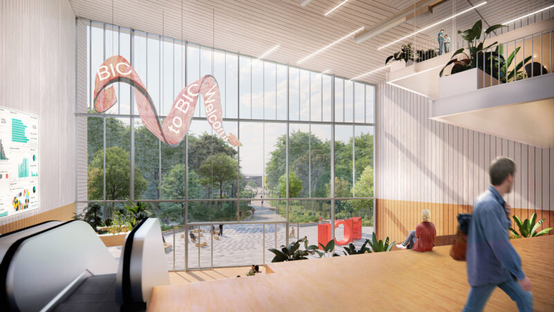 Modern lobby with large windows, indoor plants, and a welcome sign. People and green trees outside.